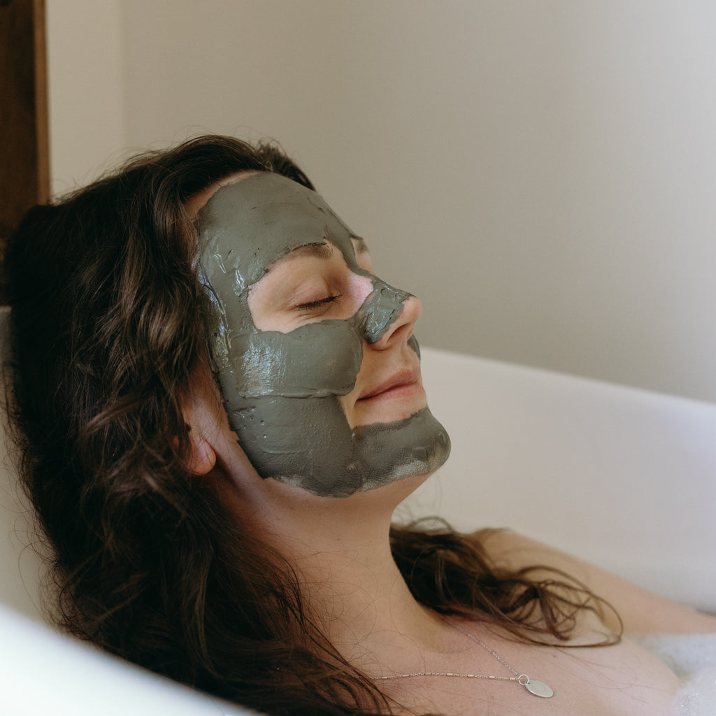 Woman relaxing in a bathtub with Oh Canada Gifts Ancient Mud Facial Mask applied to her face for deep pore cleansing.
