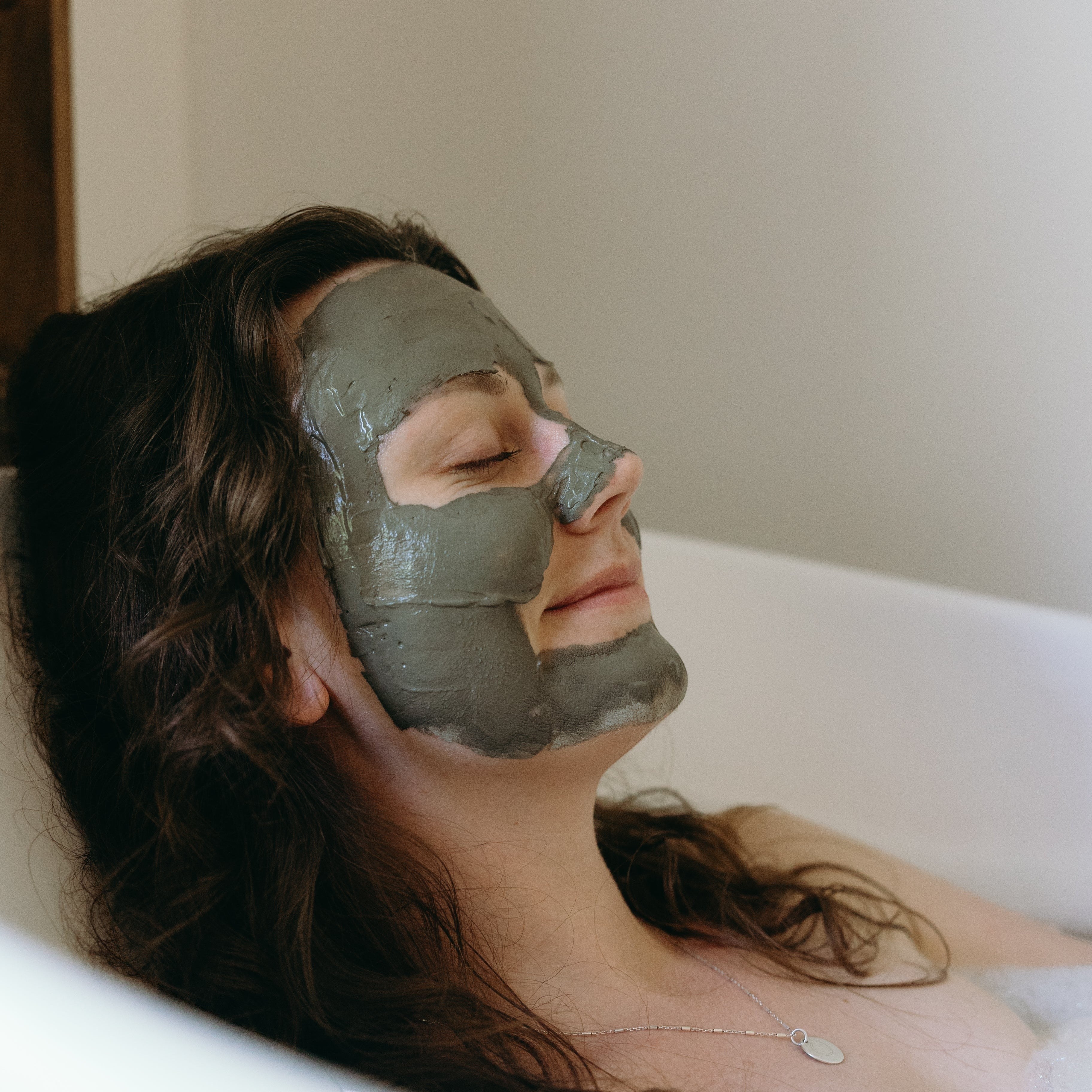 Woman relaxing in a bathtub with Oh Canada Gifts Ancient Mud Facial Mask applied to her face for deep pore cleansing.
