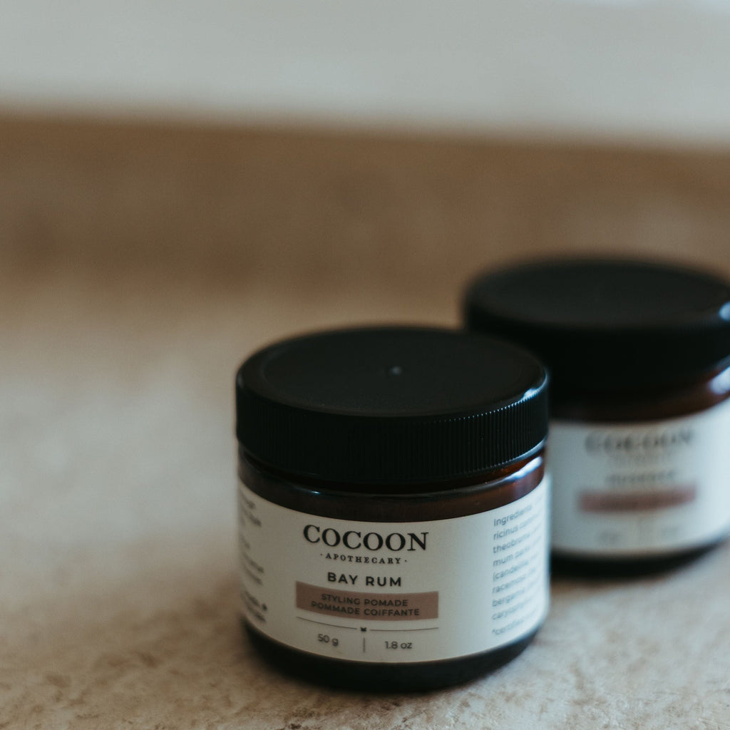 Oh Canada Gifts Cocoon Apothecary Bay Rum styling pomade in amber glass jars on a neutral textured surface.
