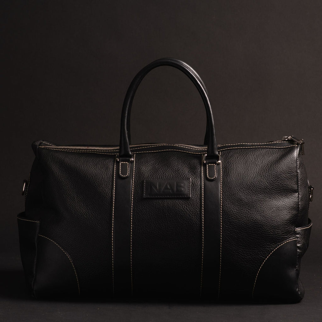 Oh Canada Gifts NAB black full-grain leather duffle bag with contrast stitching and top handles on a dark background.