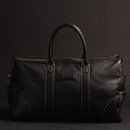 Oh Canada Gifts NAB black full-grain leather duffle bag with contrast stitching and top handles on a dark background.