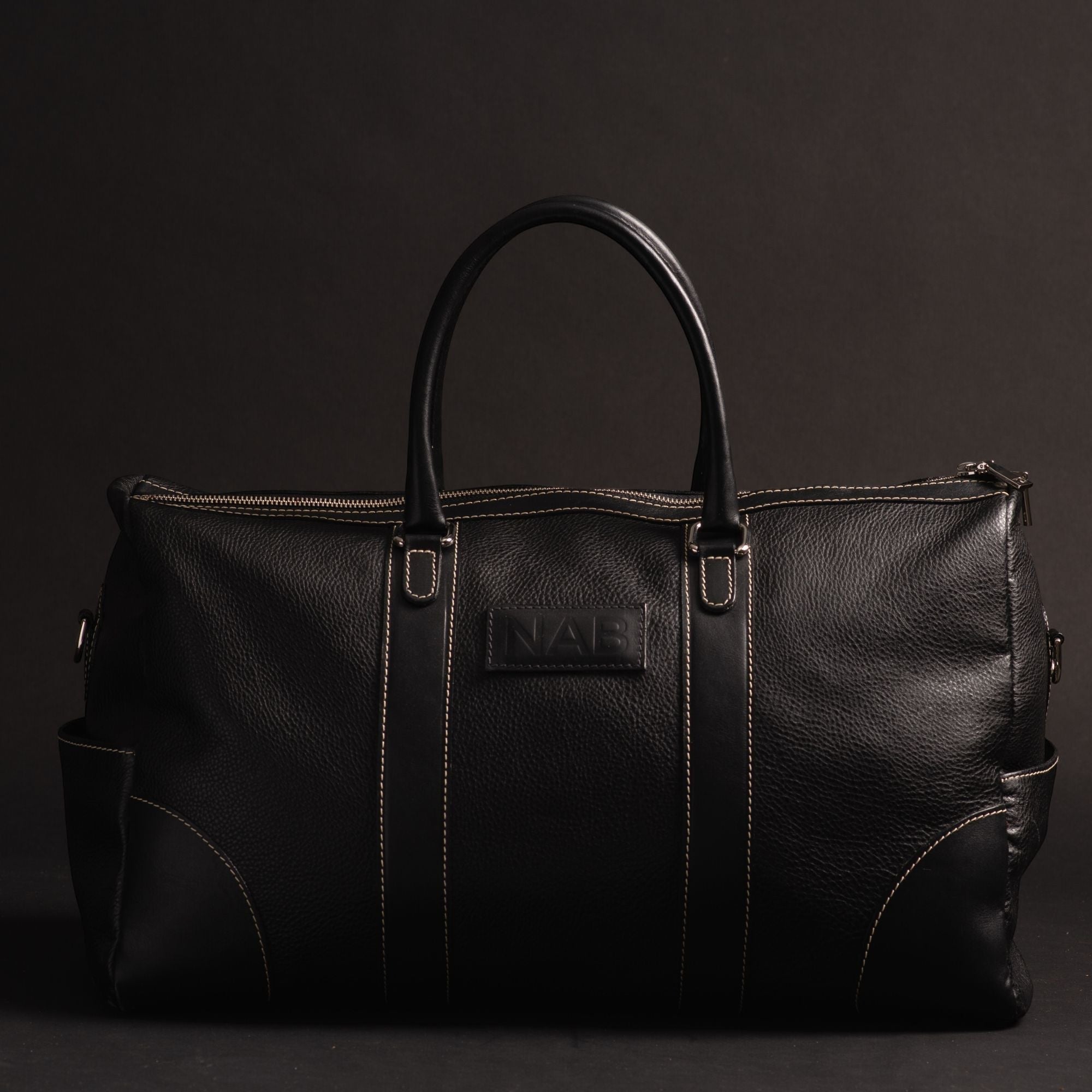 Oh Canada Gifts NAB black full-grain leather duffle bag with contrast stitching and top handles on a dark background.