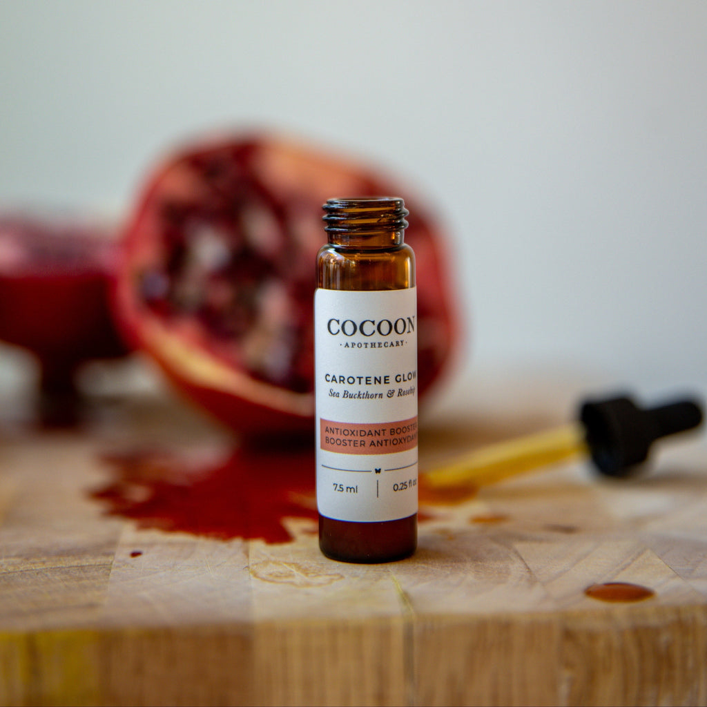 Oh Canada Gifts Carotene Glow facial serum in an amber glass bottle with a dropper, set against a sliced pomegranate.