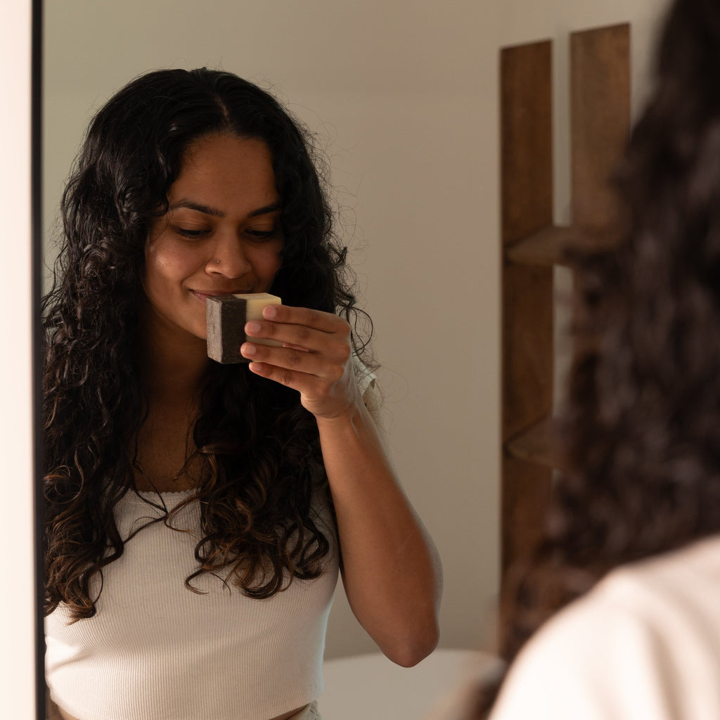 Oh Canada Gifts coffee scrub bar soap held by a woman with long curly hair smelling the invigorating scent in a mirror
