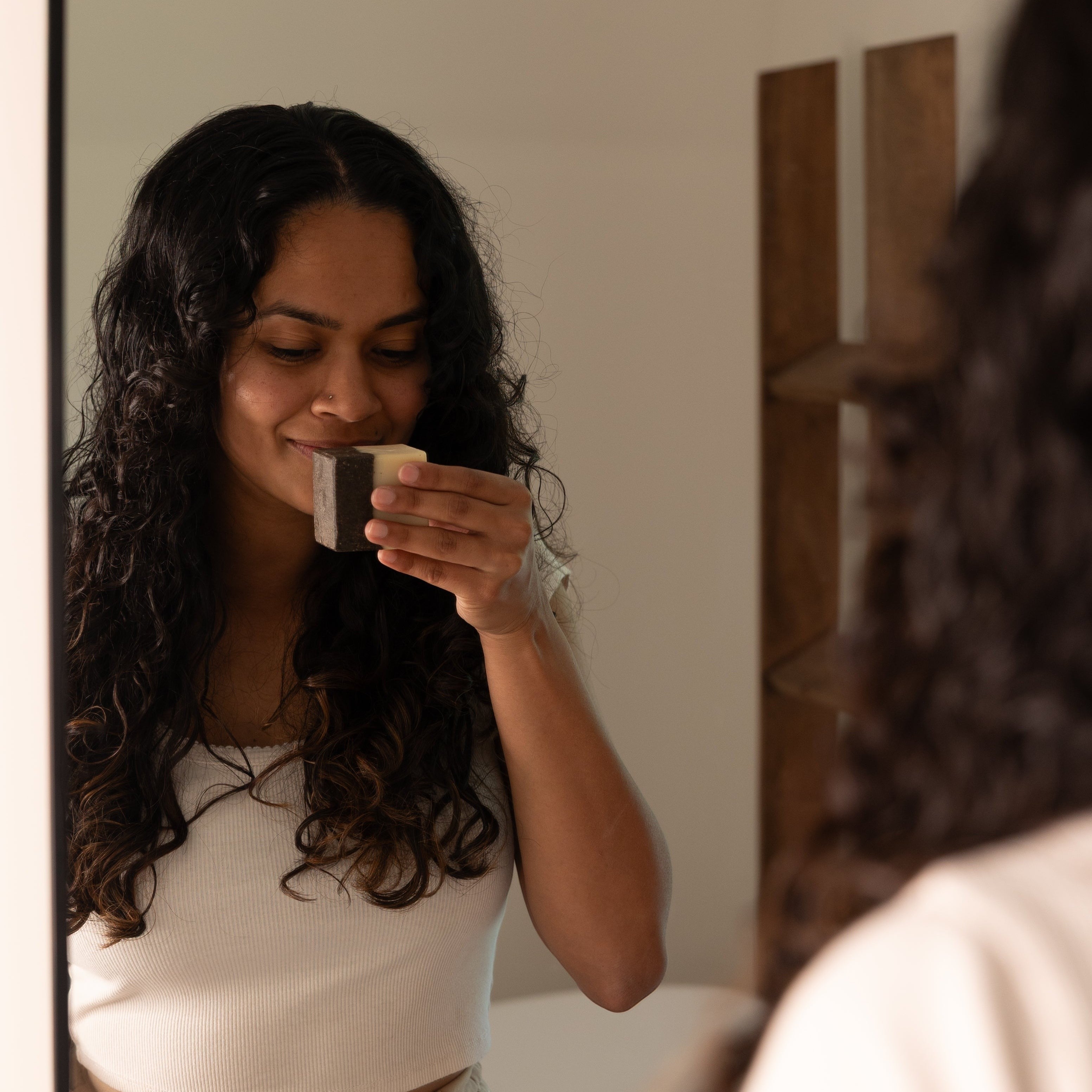 Oh Canada Gifts coffee scrub bar soap held by a woman with long curly hair smelling the invigorating scent in a mirror