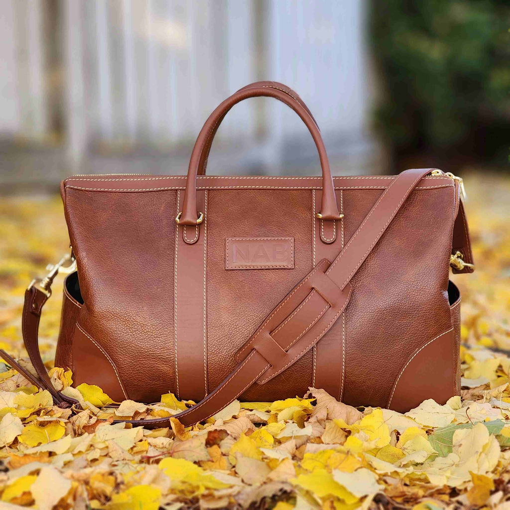 Oh Canada Gifts Shield Duffle bag in cognac full-grain leather with shoulder strap, resting on autumn leaves.