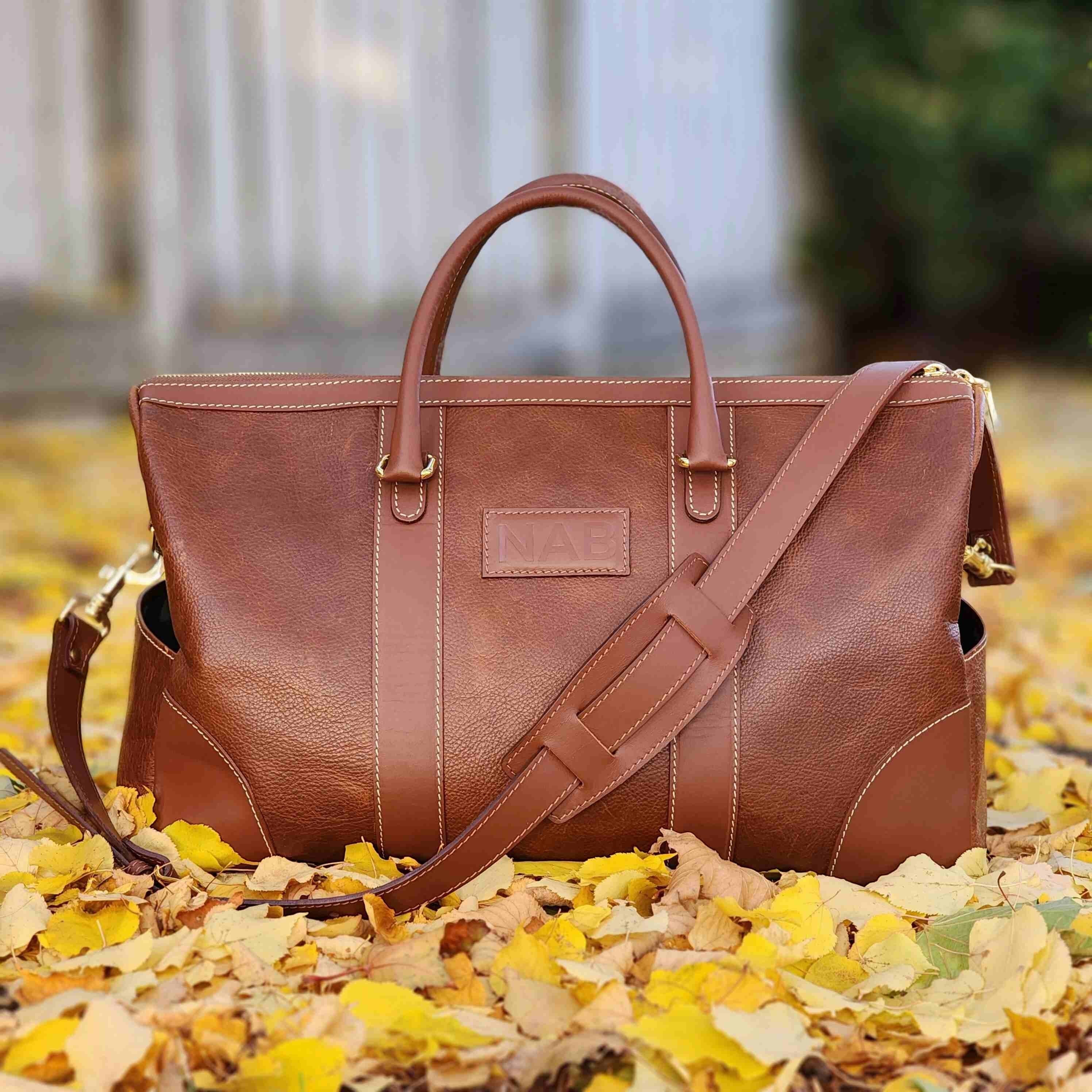 Oh Canada Gifts Shield Duffle bag in cognac full-grain leather with shoulder strap, resting on autumn leaves.