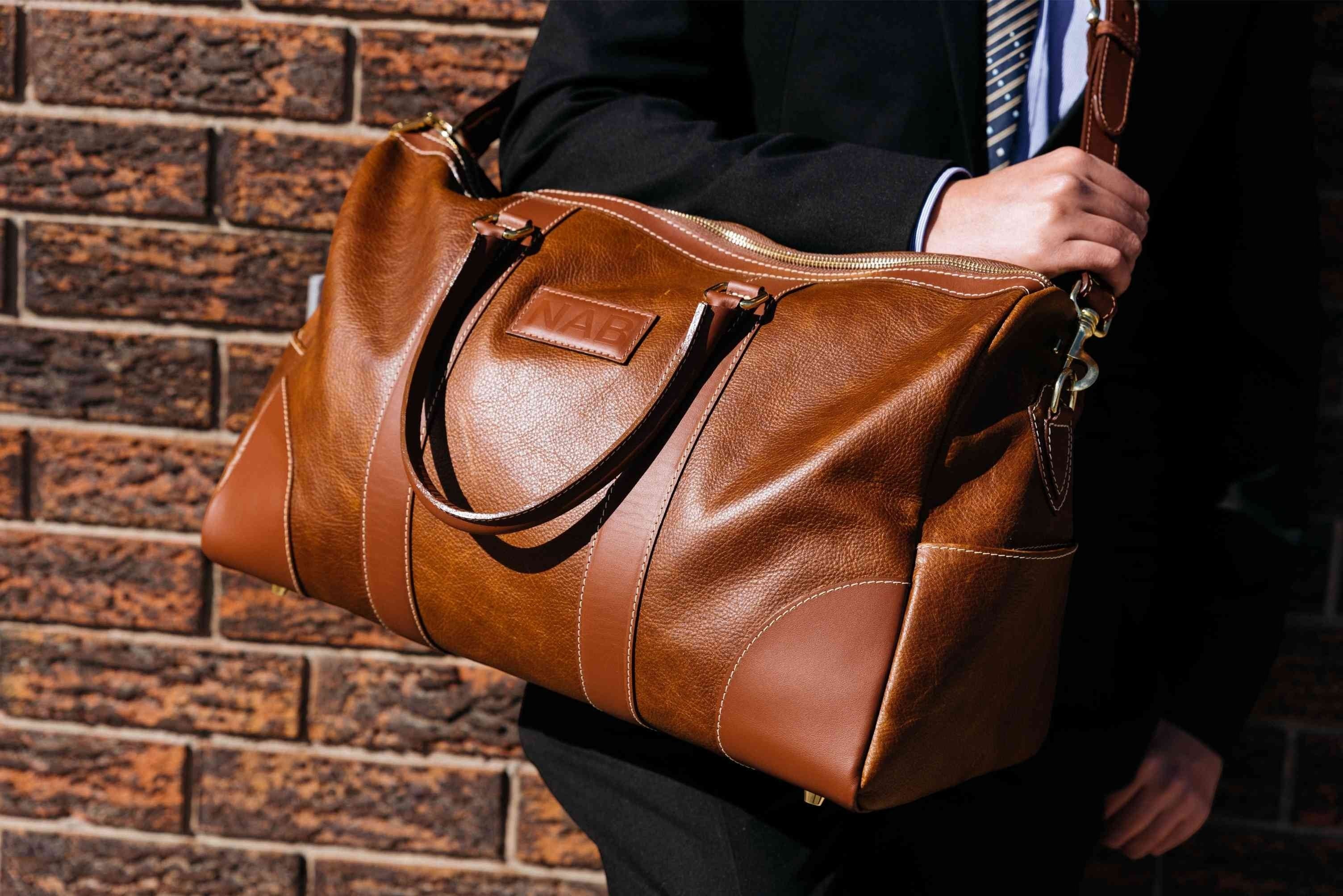 Oh Canada Gifts NAB Leather cognac full-grain leather duffle bag held by a person in a suit against a brick wall.