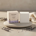Oh Canada Gifts Lavandin bath cube in beige packaging next to a solid white bath cube on a stone with lavender sprigs.