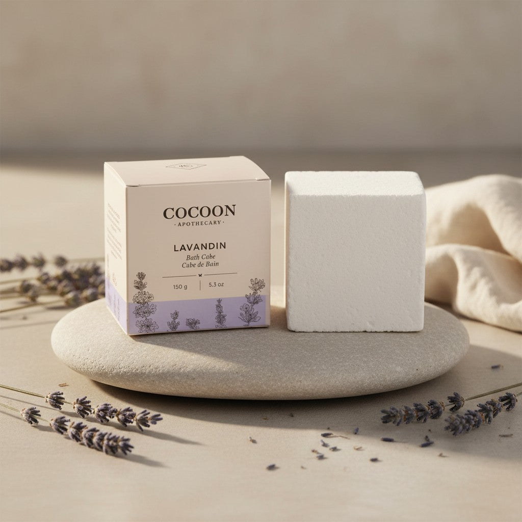 Oh Canada Gifts Lavandin bath cube in beige packaging next to a solid white bath cube on a stone with lavender sprigs.