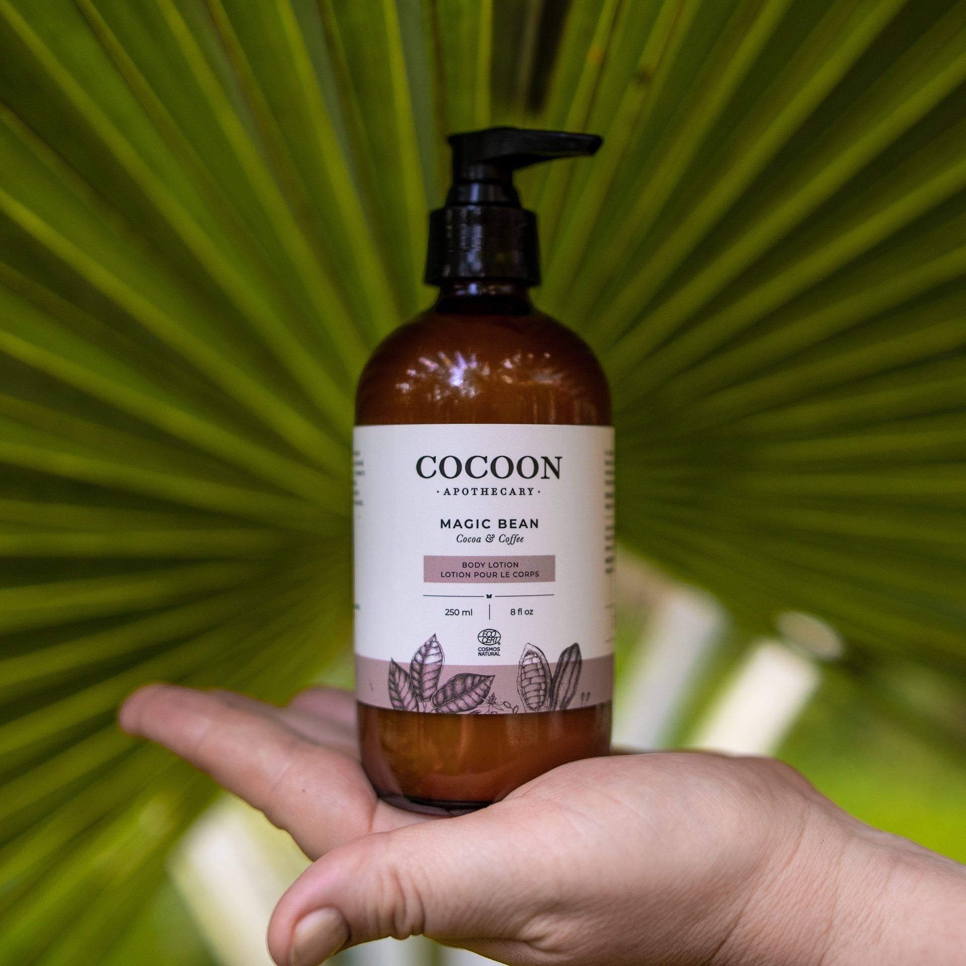 Oh Canada Gifts Magic Bean body lotion in a brown glass pump bottle held in a hand against a palm leaf background.