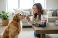 Oh Canada Gifts owner feeding a natural air-dried rabbit and beaver dog treat stick to a golden retriever at home.