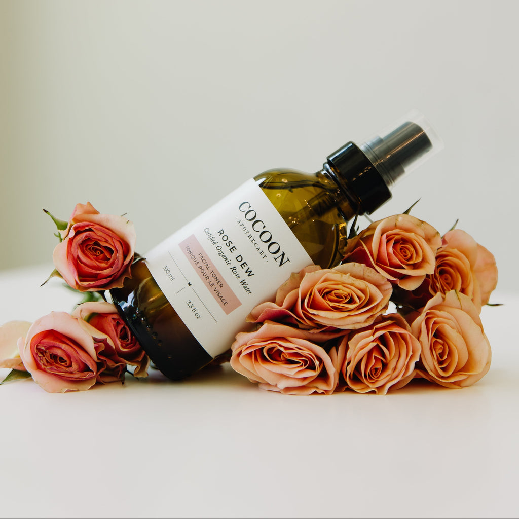 Oh Canada Gifts Rose Dew facial toner in a glass spray bottle surrounded by fresh pink roses on a white surface.
