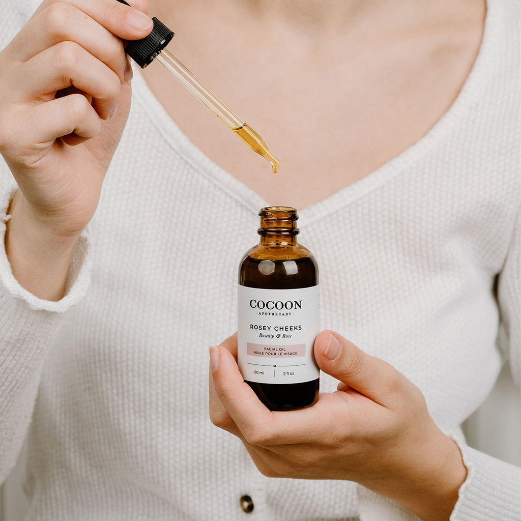 Oh Canada Gifts Rosey Cheeks facial oil in a dark glass bottle with a dropper being applied by a woman in a white top.
