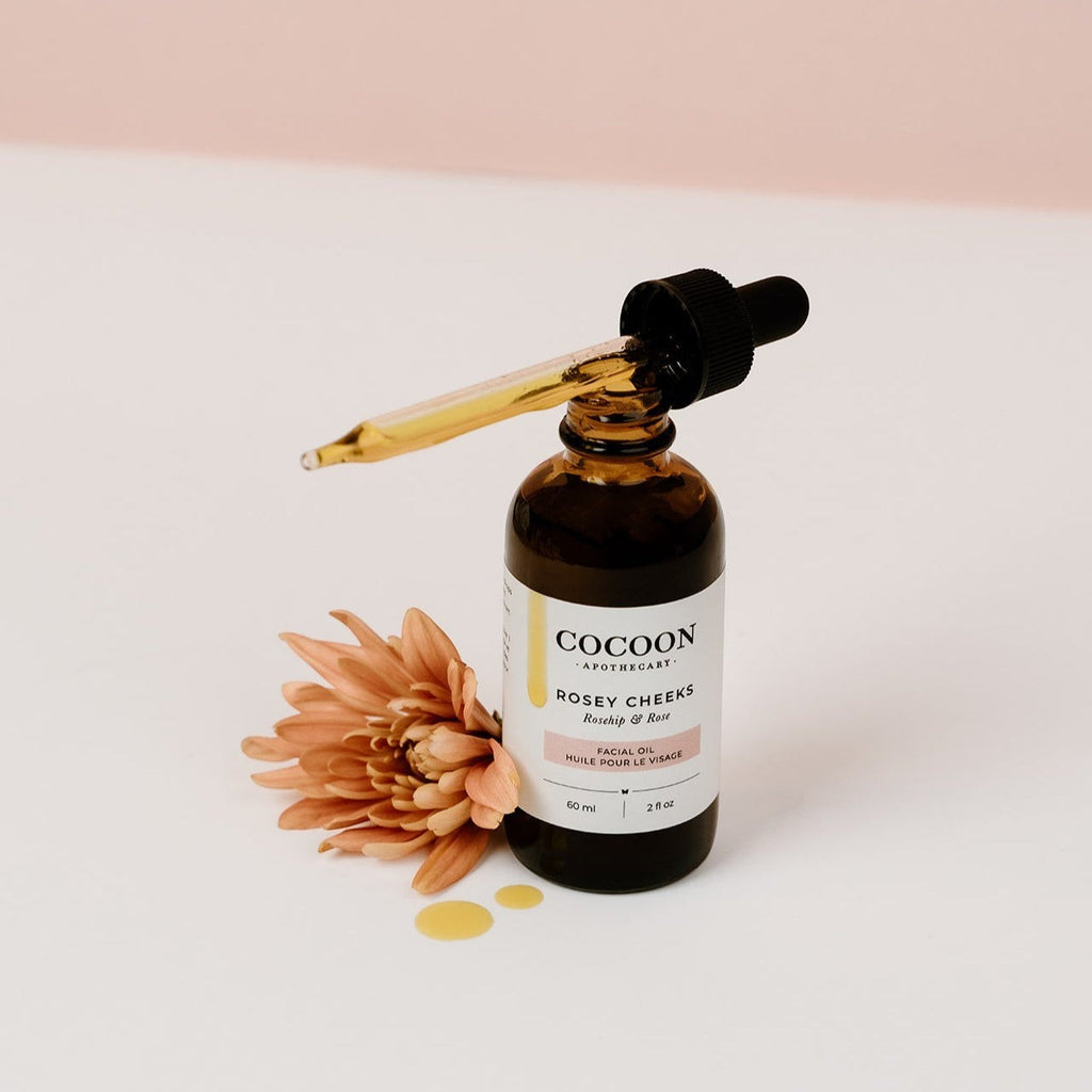 Oh Canada Gifts Rosey Cheeks facial oil in an amber glass dropper bottle with a flower on a white surface.