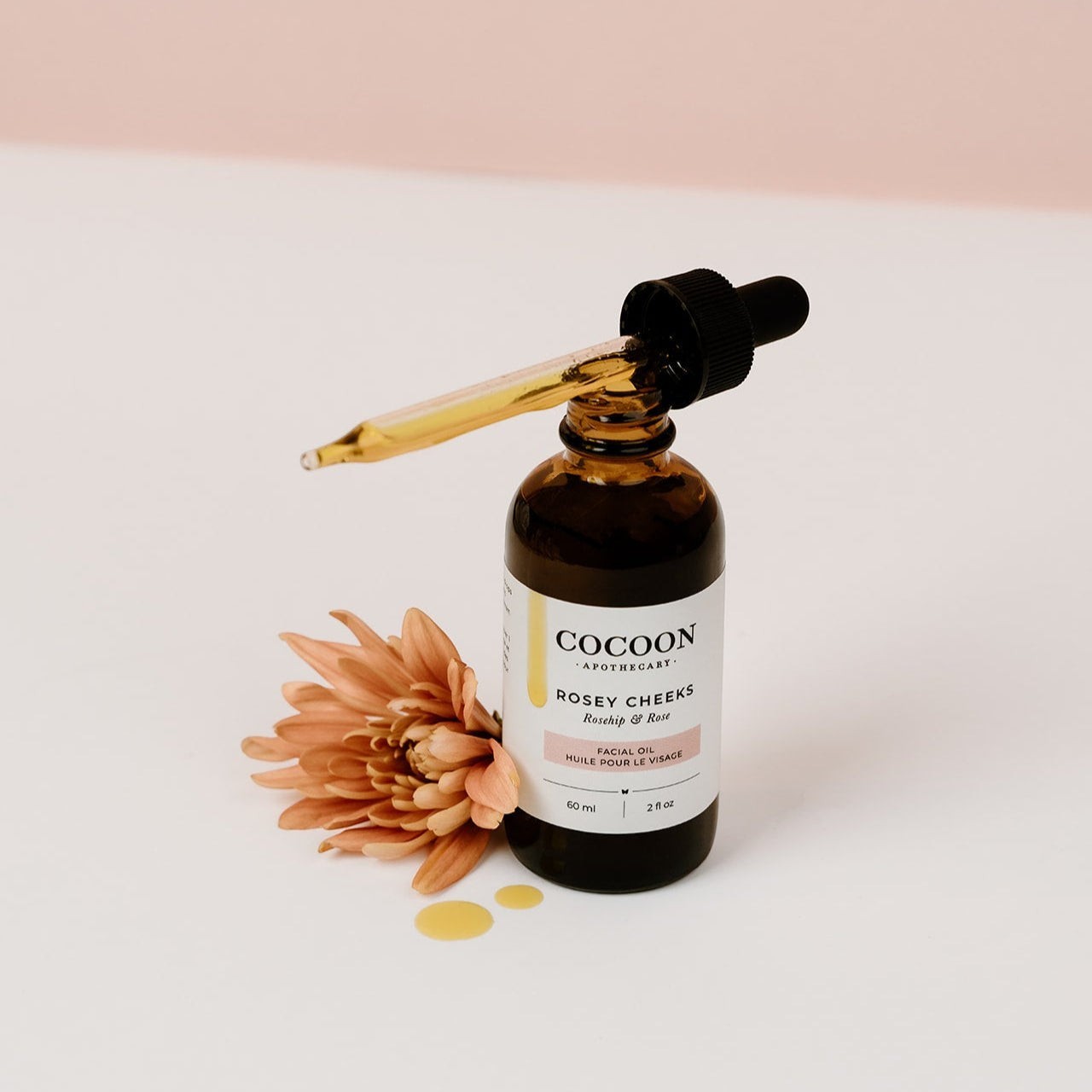 Oh Canada Gifts Rosey Cheeks facial oil in an amber glass dropper bottle with a flower on a white surface.