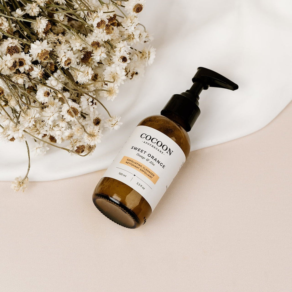 Oh Canada Gifts Sweet Orange Exfoliating Cleanser in a brown glass pump bottle next to dried white flowers.