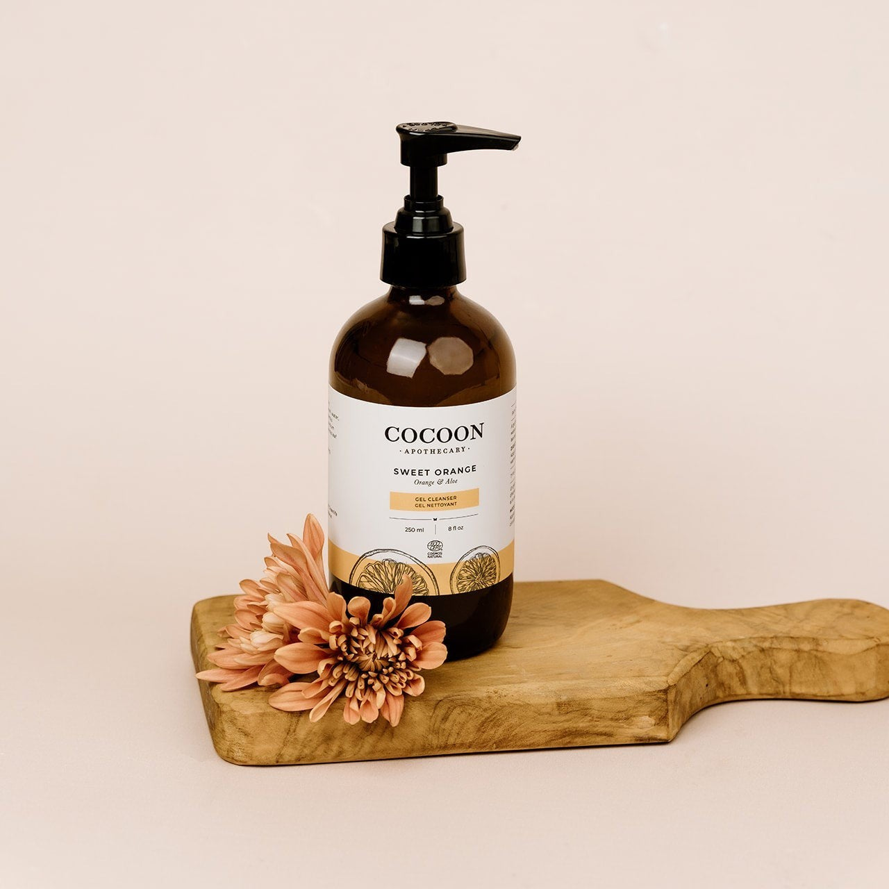 Oh Canada Gifts Sweet Orange Gel Cleanser in an amber bottle with a pump, displayed on a wooden board with flowers.