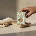Oh Canada Gifts tobacco leaf soap bar in a green box on a wooden soap dish with a beige cloth in a minimalist setting.