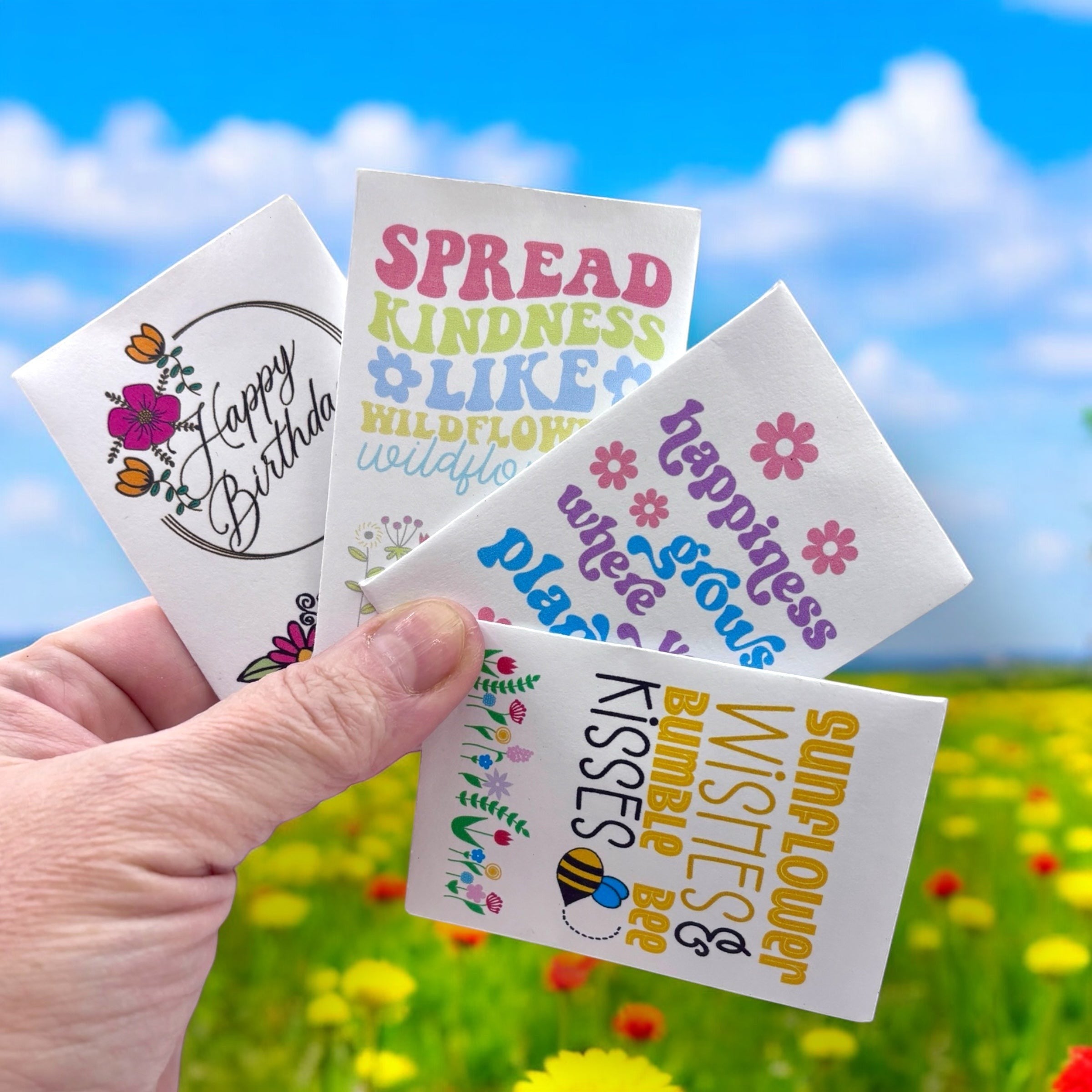 Oh Canada Gifts wildflower seed packets with colorful floral designs and inspirational text held against a meadow.