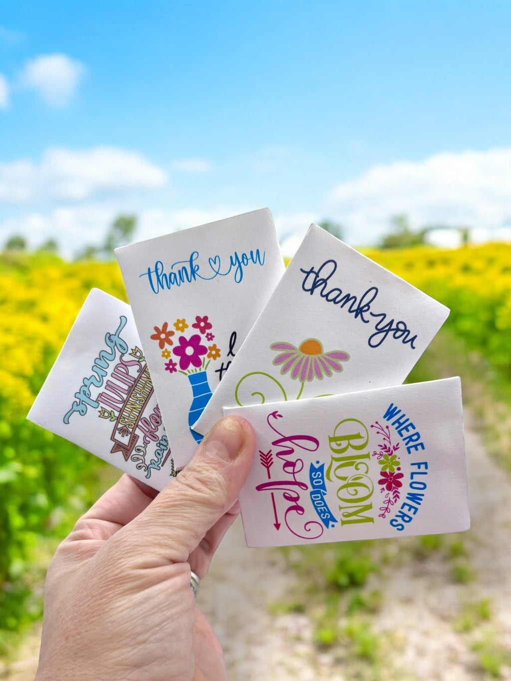 Oh Canada Gifts wildflower seed packets with colorful floral designs and thank you messages held in a hand outdoors.
